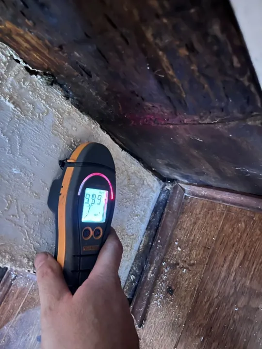 A moisture meter held against damp plaster beneath a dark timber beam